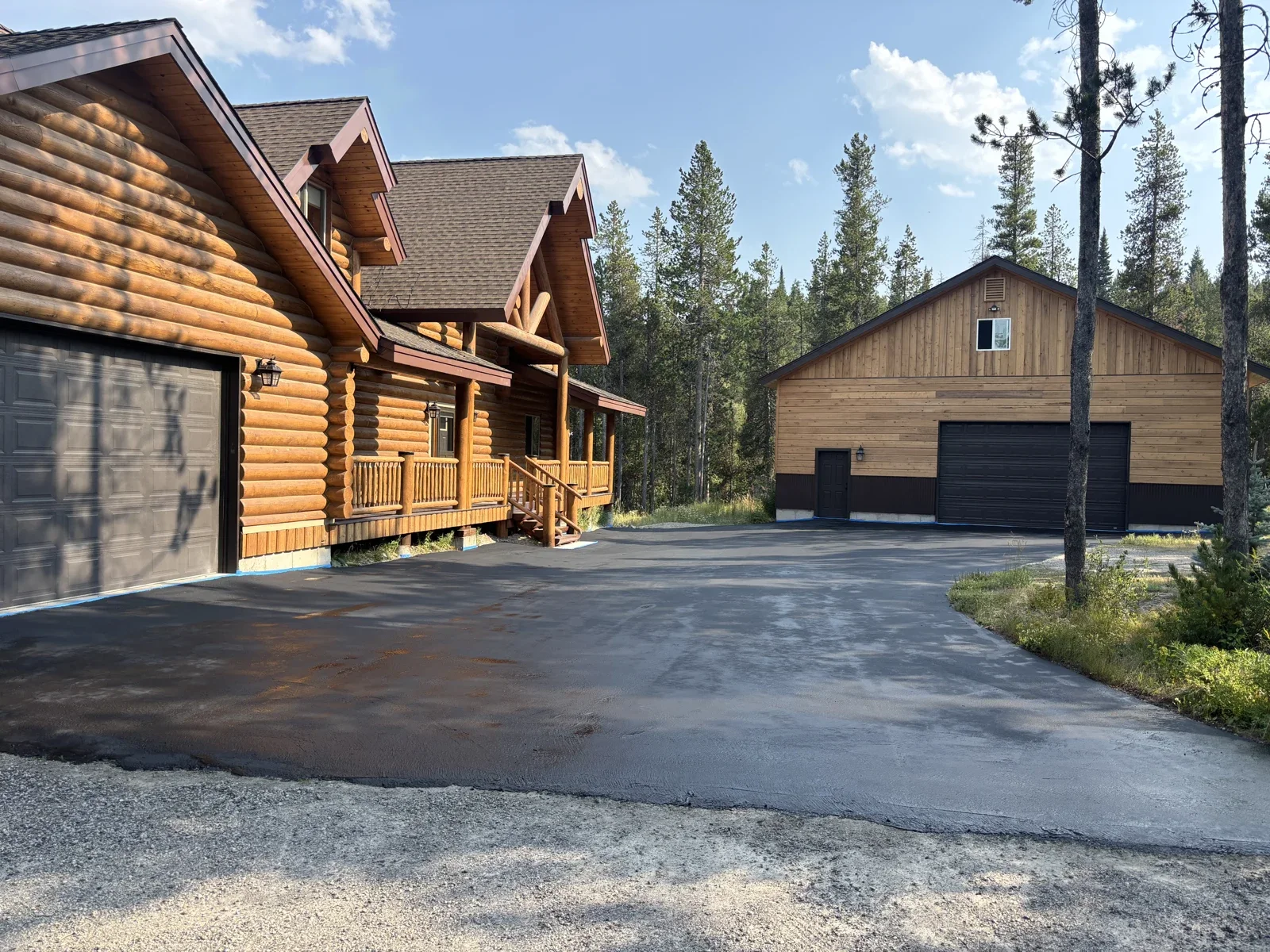 Asphalt driveway beside a log home and detached garage, surrounded by pine trees.